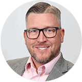 Patrick Rosen, VP of Marketing, Panorama Eyecare | Caucasian male is shown wearing a grey suit, pink button up shirt and black glasses sitting against a grey background.